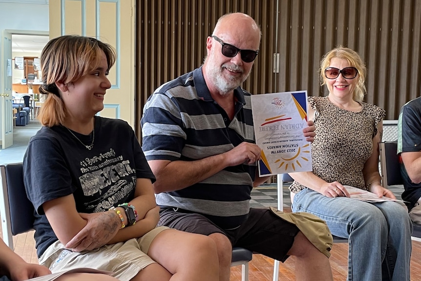 A bald man wearing dark glasses holding a certificate upside down and smiling while sitting between two people in chairs.