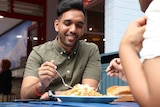 A mid-shot of a man smiling as he sits at an outdoor table eating food from a  plate.