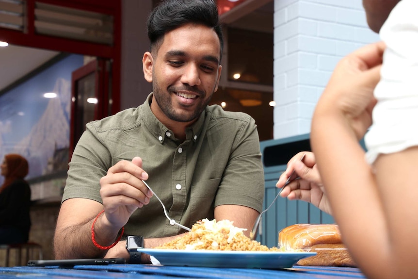 A mid-shot of a man smiling as he sits at an outdoor table eating food from a  plate.