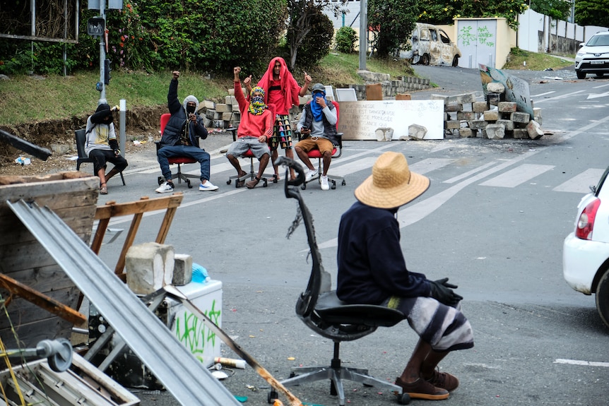 A group of people sit on chairs with their fists in the air surrounded by makeshift roadblocks made from debris in the street