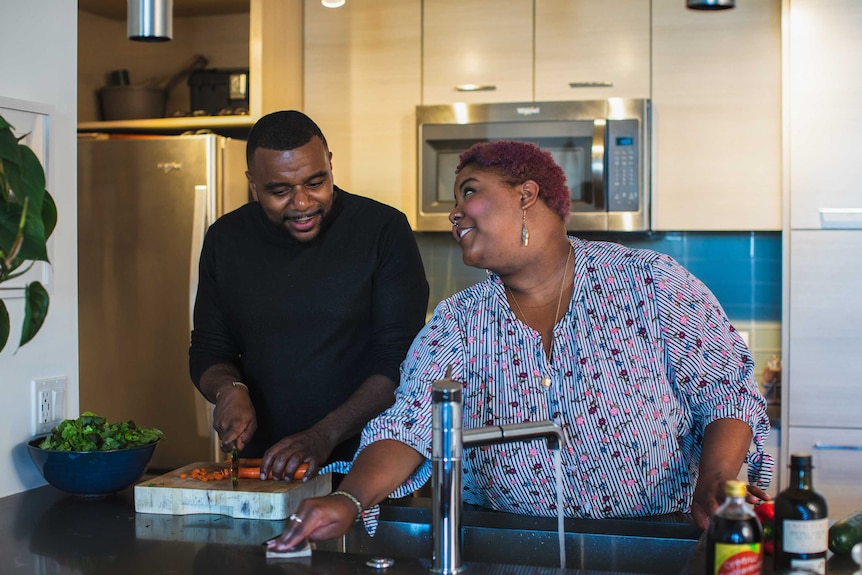 A man is chopping carrots next to a woman wiping down a sink and they are looking at each other