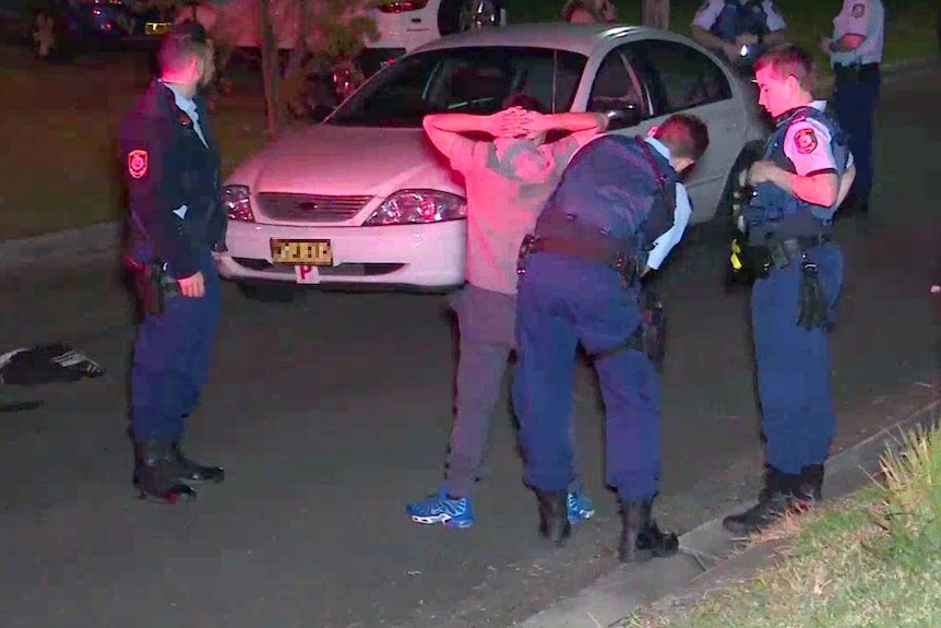 Three police search a man with his hands on his head in front of a car.