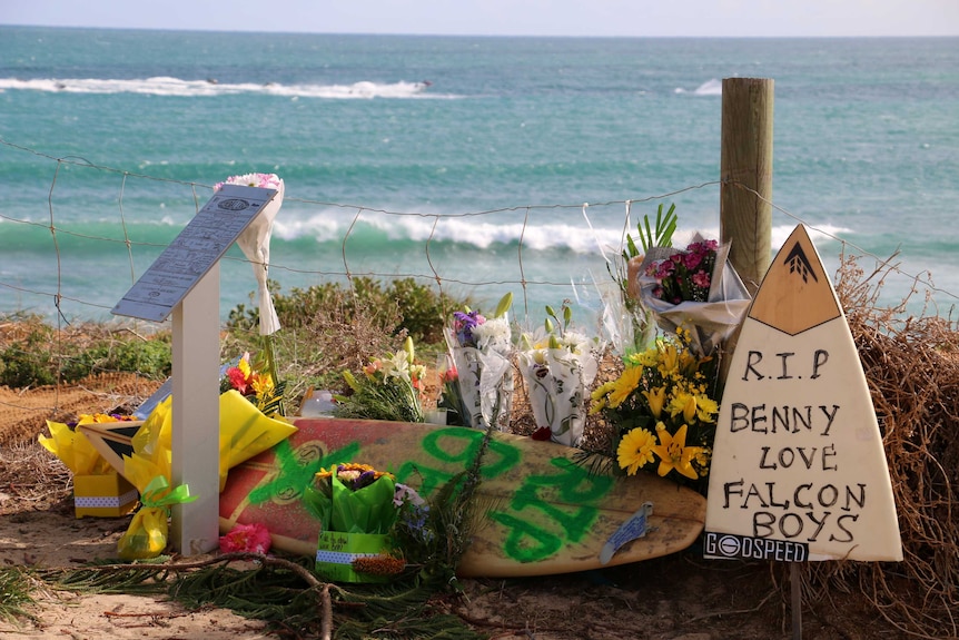 Tributes including flowers and surfboards rest up against a wire fence in front of Falcon beach, with waves in the background.