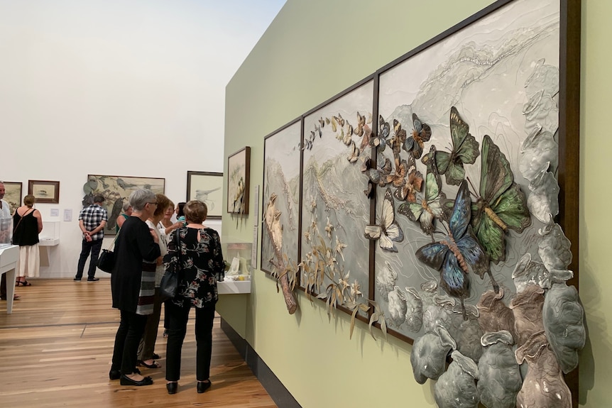 People looking at a piece of art where butterflies emerge from the canvas.
