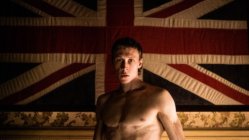 A topless man with short cropped hair stands in front of large Union Jack flag hanging from ornate patterned wallpaper.