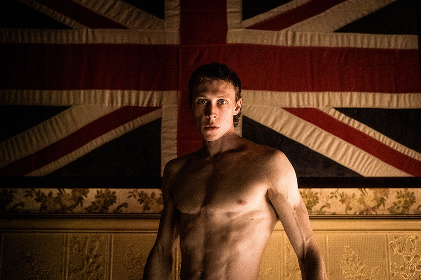 A topless man with short cropped hair stands in front of large Union Jack flag hanging from ornate patterned wallpaper.