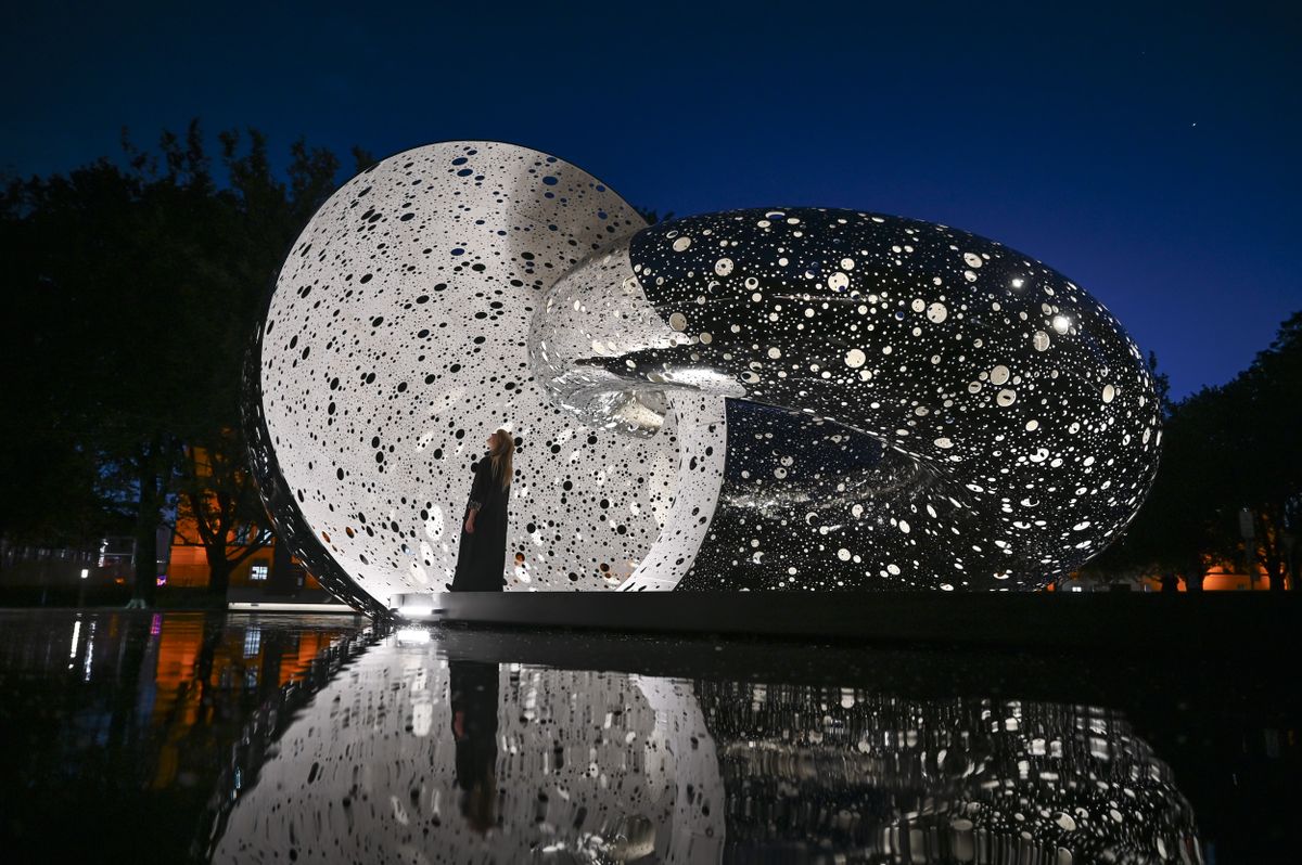 A woman stands in the mouth of a metal statue full of holes emitting light beams at night.