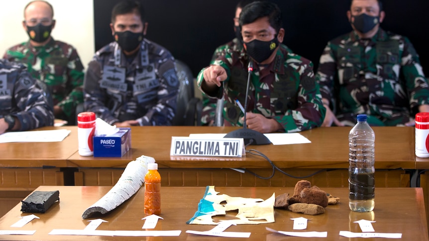 Men in navy attire sit together. One points at items from a submarine in the foreground.