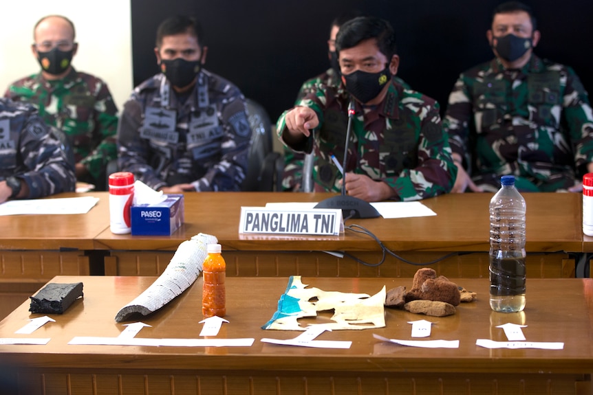 Men in navy attire sit together. One points at items from a submarine in the foreground.