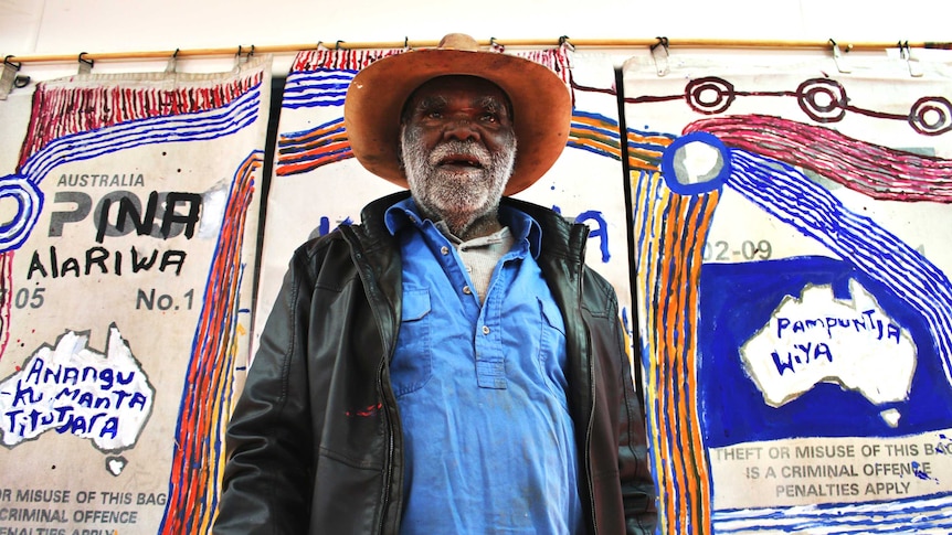 Colour photograph of artist Mumu Mike Williams standing in front of his artwork, painted onto old Australian Post mailbags