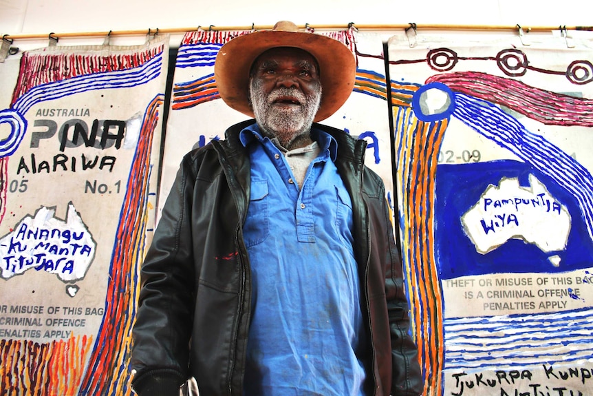 Colour photograph of artist Mumu Mike Williams standing in front of his artwork, painted onto old Australian Post mailbags