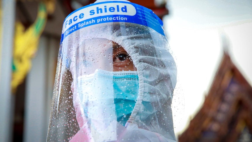 A man in a mask, face shield and PPE stands in front of a Thai temple 