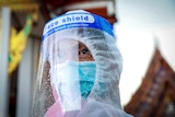A man in a mask, face shield and PPE stands in front of a Thai temple 
