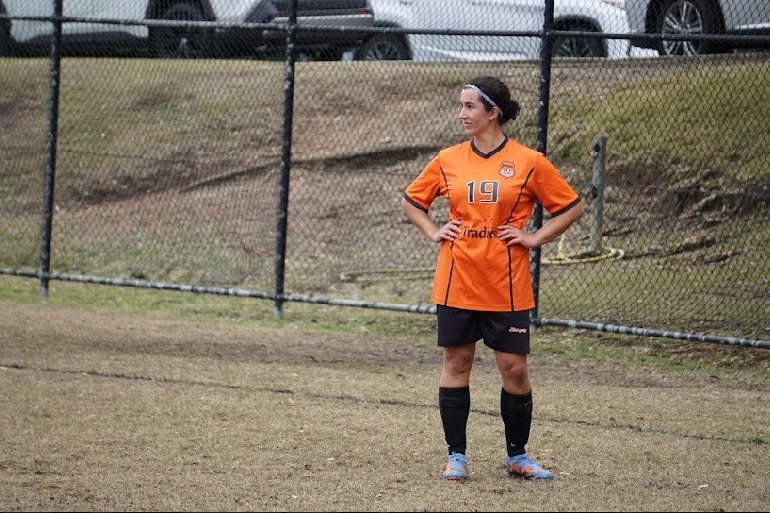 A woman in orange soccer gear watches the game unfold.