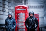 A man and a woman bundled up in winter gear walk through snow with red telephone boxes behind them