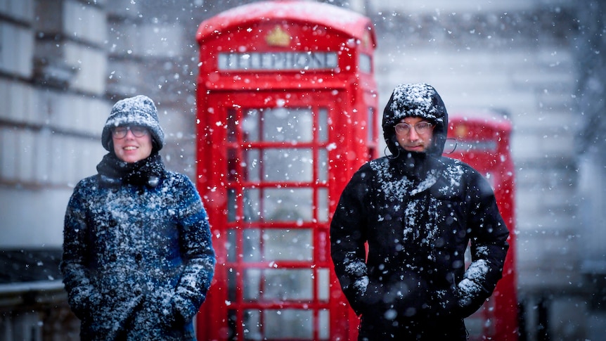 A man and a woman bundled up in winter gear walk through snow with red telephone boxes behind them