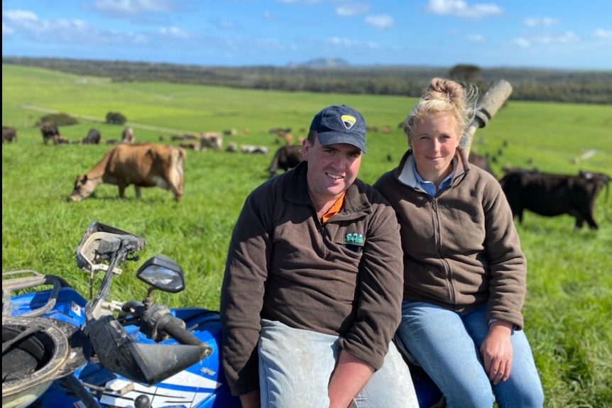 Young dairy farmers Ryan and Brighid Langley sit on a quad bike in lush green paddock filled with cows.