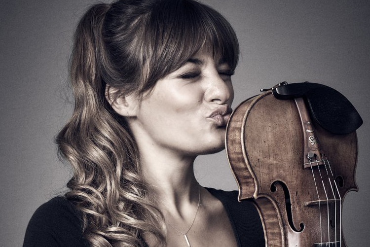 Violinist Nicola Benedetti holds a violin upside down and is pretending to kiss the bottom of her violin. Her eyes are closed.