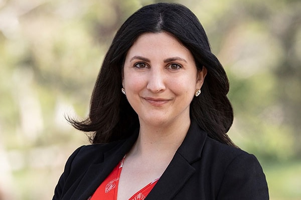 A dark-haired woman with dark eyes wearing a black jacket, red shirt and sparkling earrings