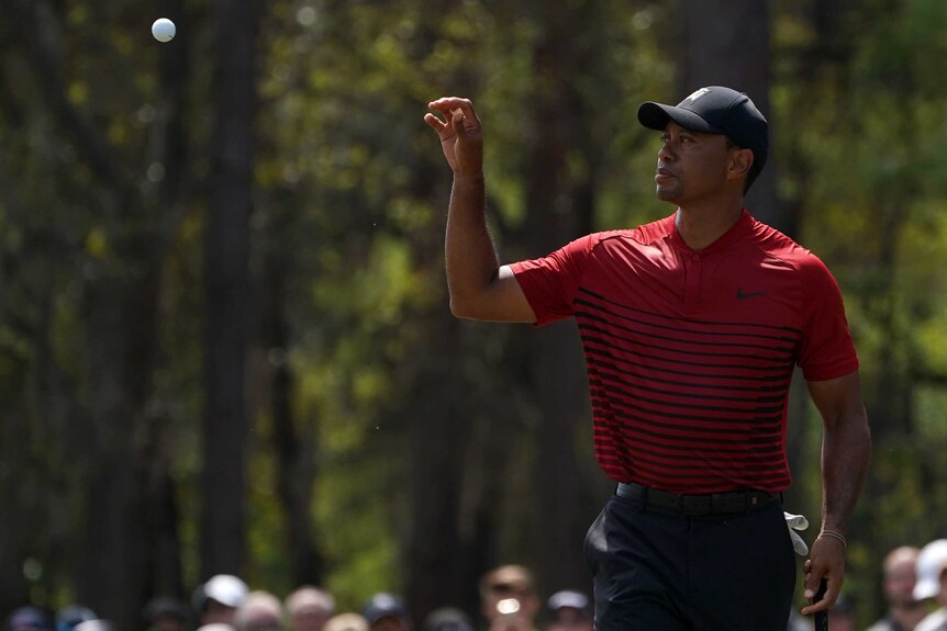Tiger Woods stands with his right hand out to catch a golf ball thrown by his caddie.