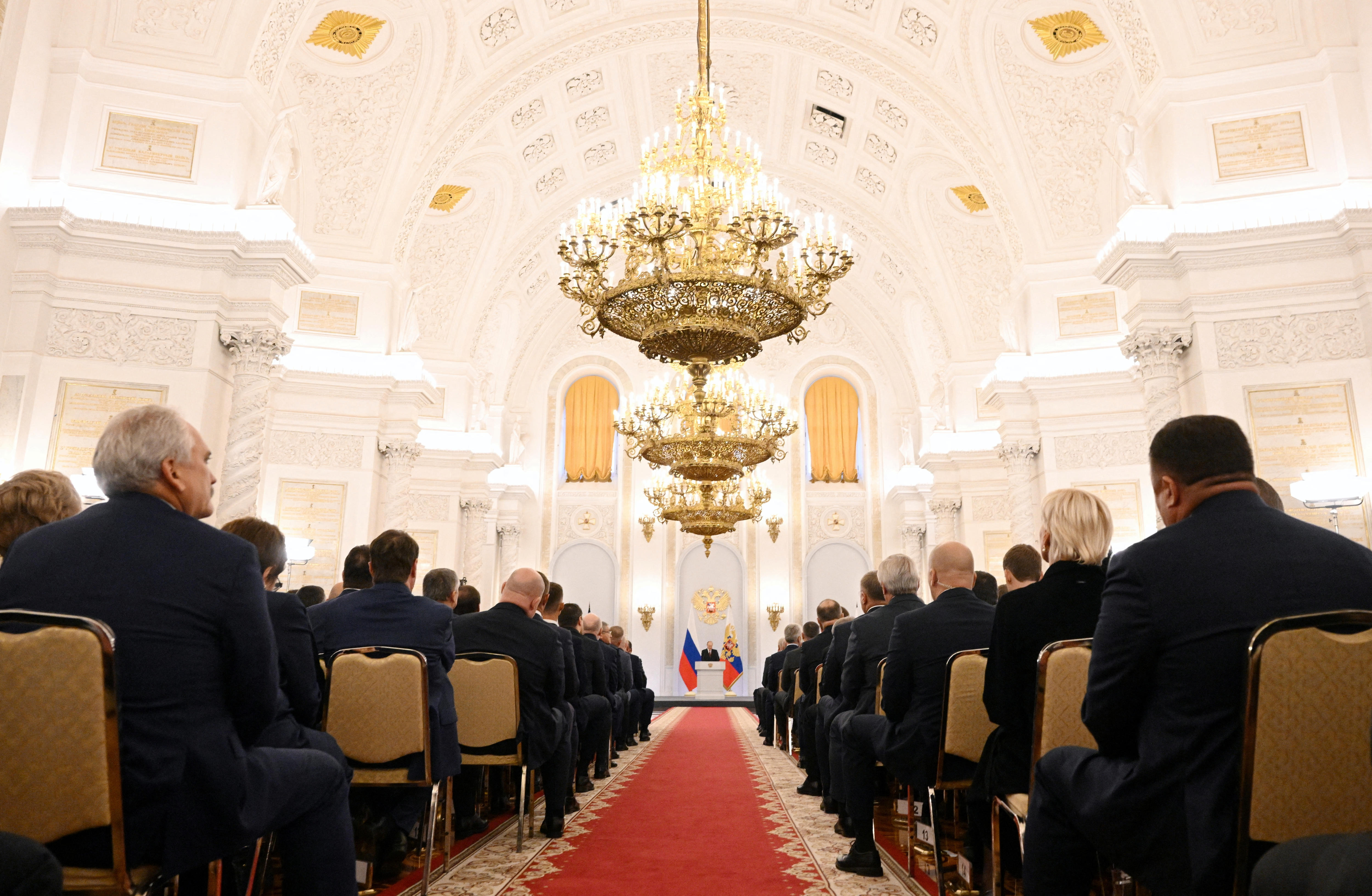 Audience members in formal attire listen to the Russian President in a grand white room.
