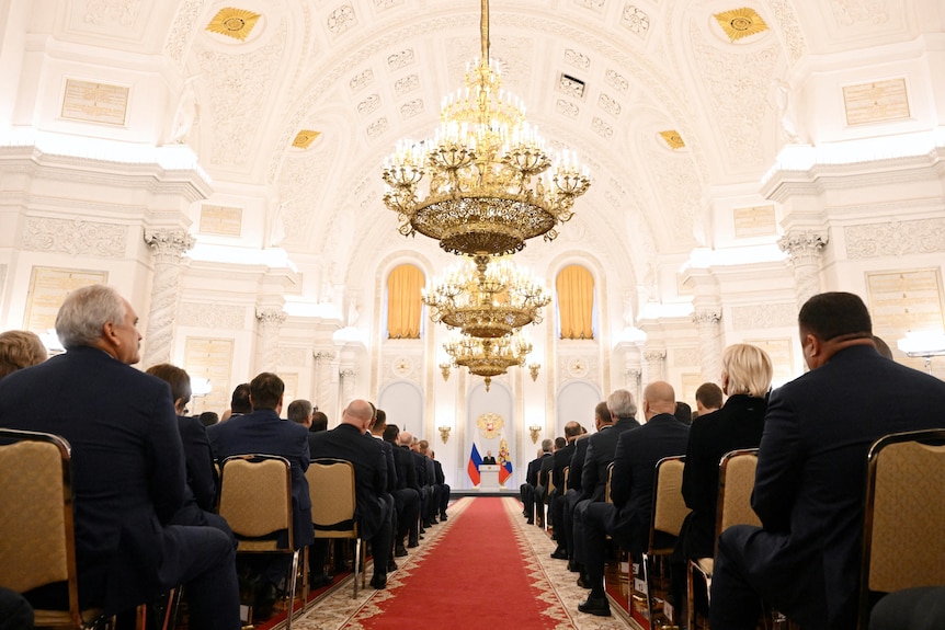 Audience members in formal attire listen to the Russian President in a grand white room.
