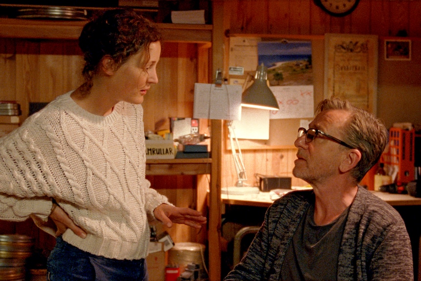 A woman in her late 30s in a white knitted jumper speaking to a man in his 60s in a wooden room, reels of films in the back