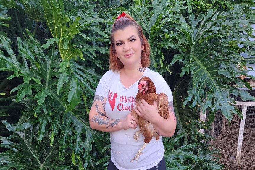 Bonney stands in front of a large philodendron and smiles as she holds a chicken while wearing a "mother clucker" t-shirt.