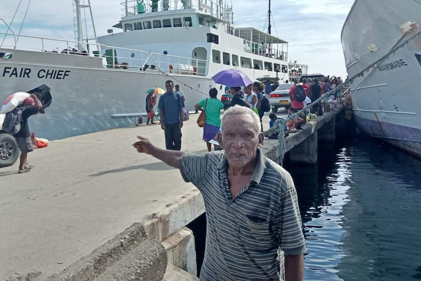 A man standing on the wharf is pointing to a ferry and there is a queue of people waiting to board the ferry behind him.