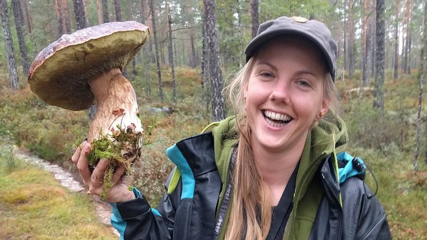 A woman wearing hiking attire smiles while holding up a comically-large mushroom.
