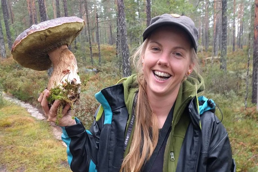 A woman wearing hiking attire smiles while holding up a comically-large mushroom.