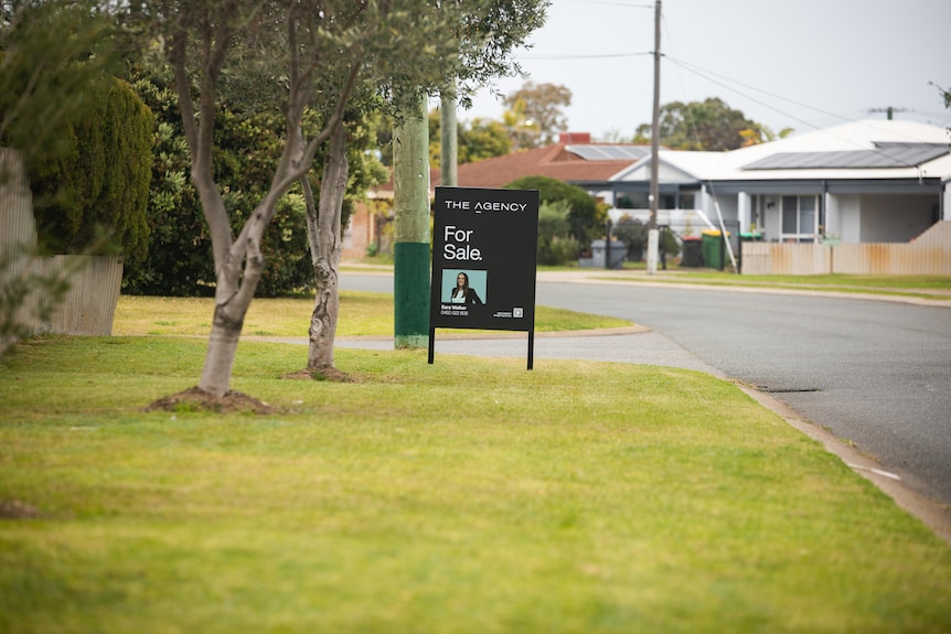 A black 'For Sale' sign on a green front lawn next to a street.