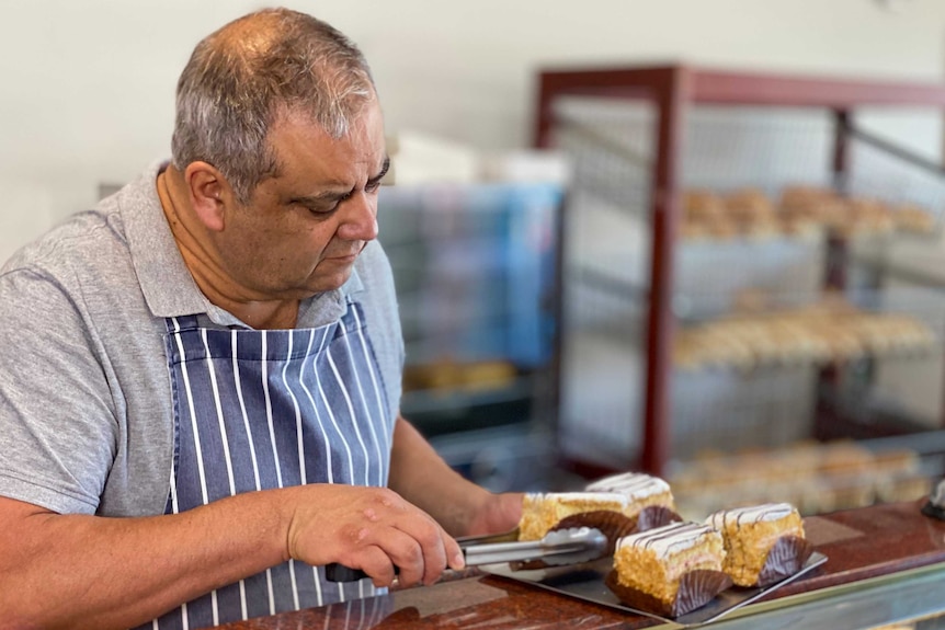 A man in a striped apron holds a cake in a pair of tongs.