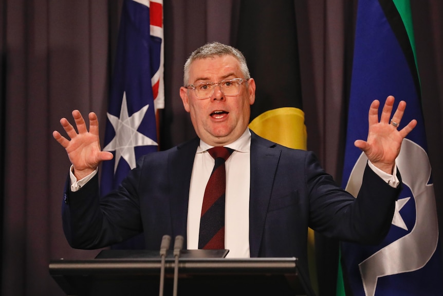 A man in a suit stands a podium holding both his hands in the air