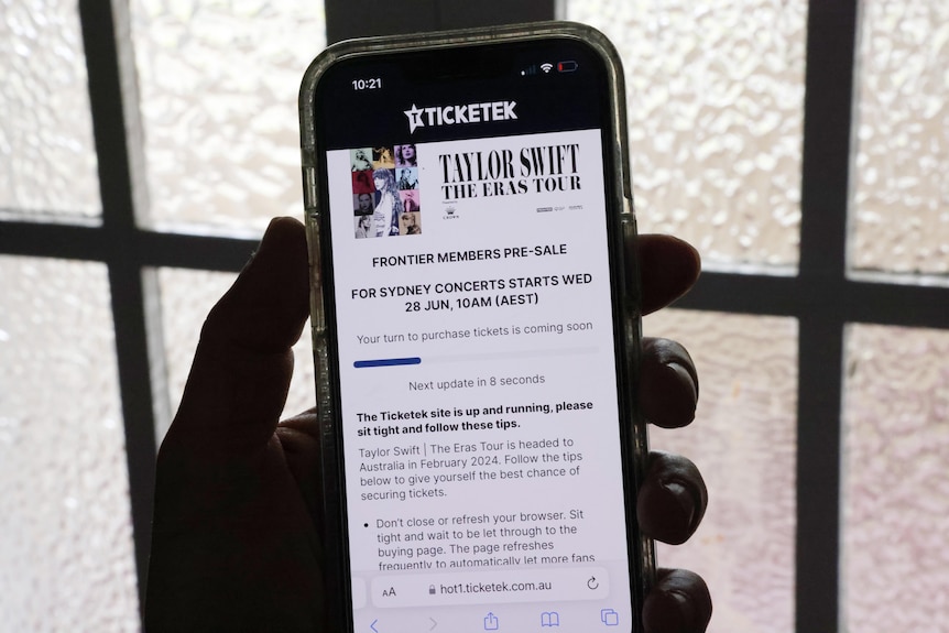 A hand holding a mobile phone in front of an opaque window, showing Taylor Swift Eras Tour Ticketek ticket queuing page