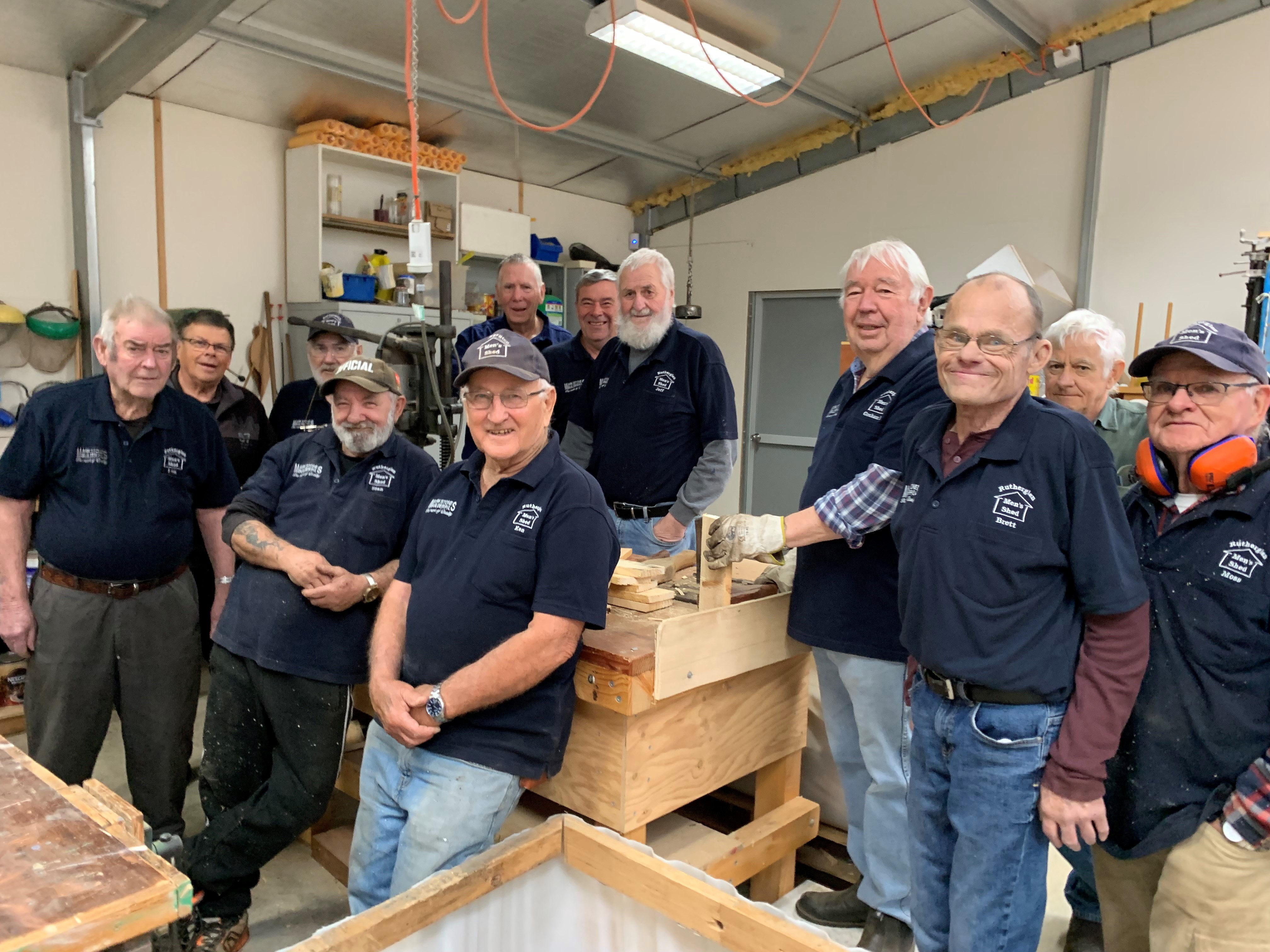 A dozen members of the Rutheglen Men's Shed stand around a work table in a shed.