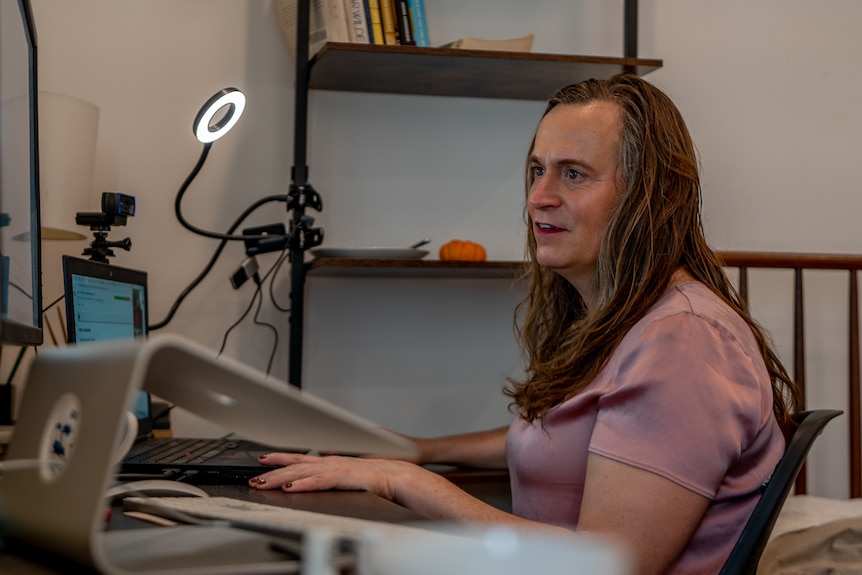 A woman in a pink shirt sits in front of a commputer desk with a ring light illuminating her face