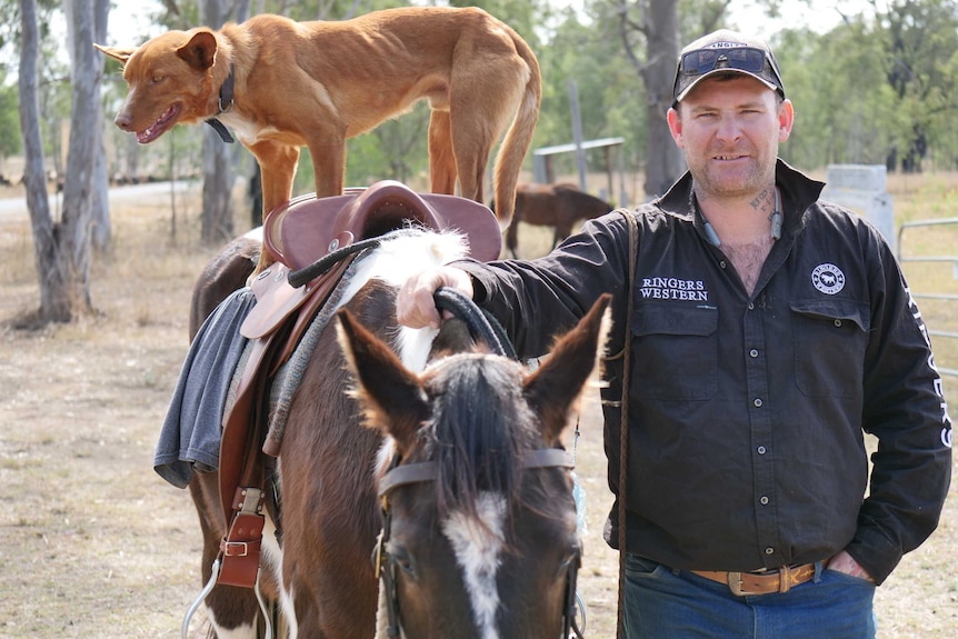 Middle aged man standing with hand on brown and white horse. A tan working dog is balancing on the horse's saddle.