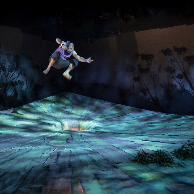 Darkened stage with a man standing on either side of it, and in the centre, above a hole, another man leaping mid-air.