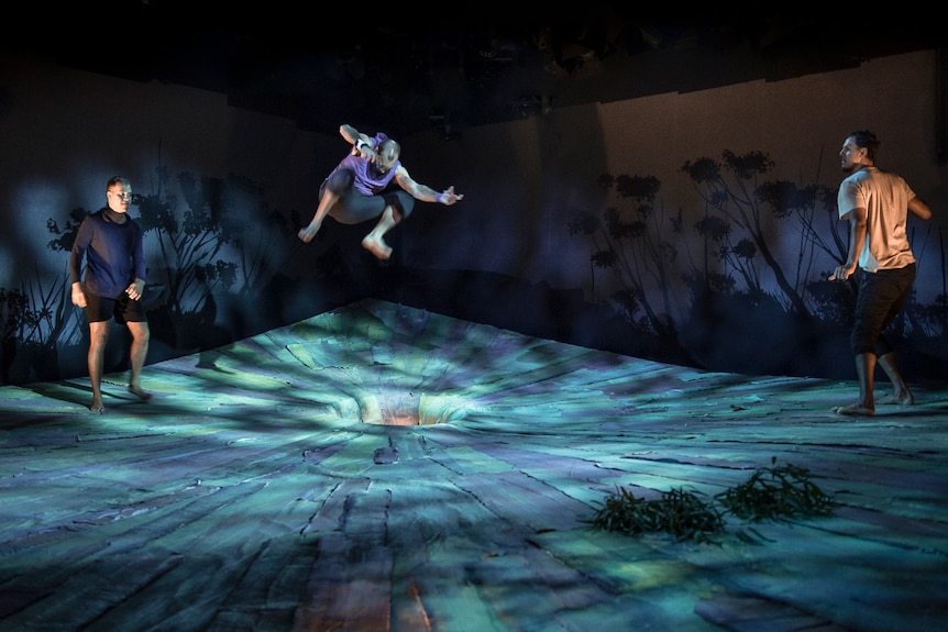Darkened stage with a man standing on either side of it, and in the centre, above a hole, another man leaping mid-air.