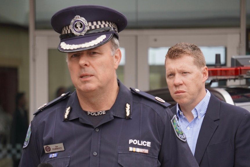 A male police officer speaks while another man wearing a blazer looks on from behind