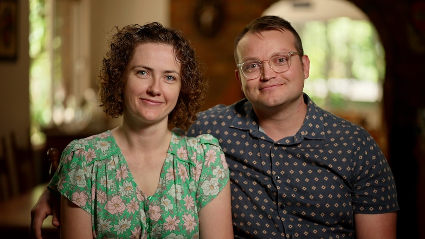 A couple sits looking directly into the camera. A man with glasses has his arm around a woman with curly hair.