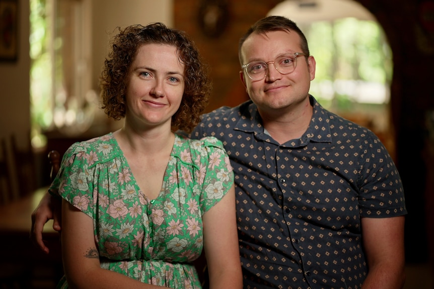 A couple sits looking directly into the camera. A man with glasses has his arm around a woman with curly hair.