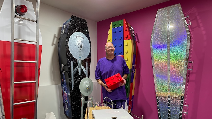 Ross stands smiling in front of coffins displayed on the wall. They are themed in Lego, glitter, a fire engine and a satellite.