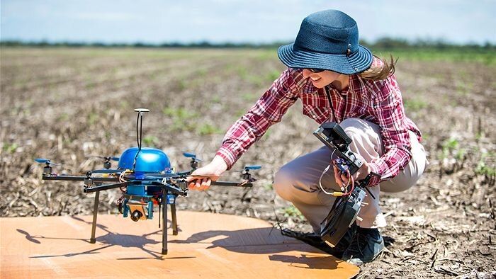 A woman in a hat crouches next to a small drone copter in a harvested field