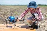 A woman in a hat crouches next to a small drone copter in a harvested field