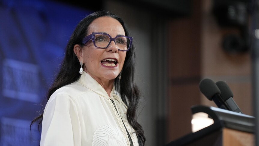 Linda Burney wearing white shirt and purple glasses standing in front a podium with a blue background