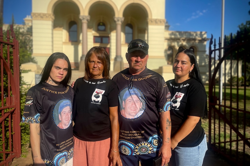 Serious man and woman stand next to two women, in-front of court house, all wearing tee-shirts with a man's photo.