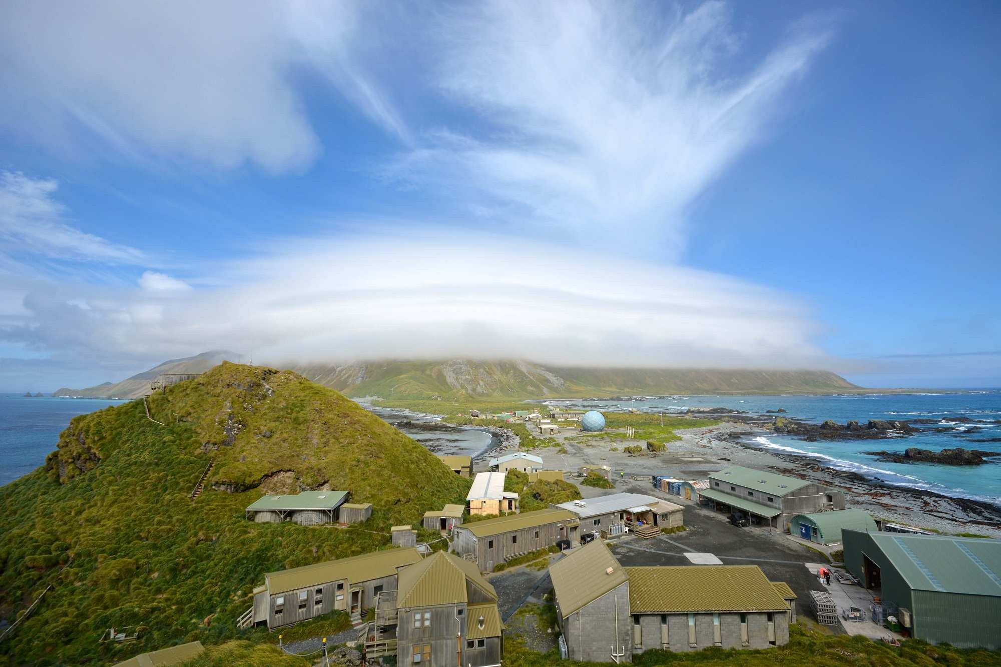 A view of buildings and small structures on an island surrounded by green mountains and blue sky.