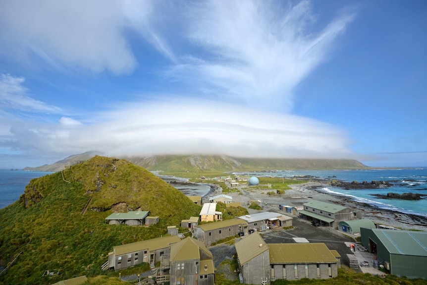 A view of buildings and small structures on an island surrounded by green mountains and blue sky.